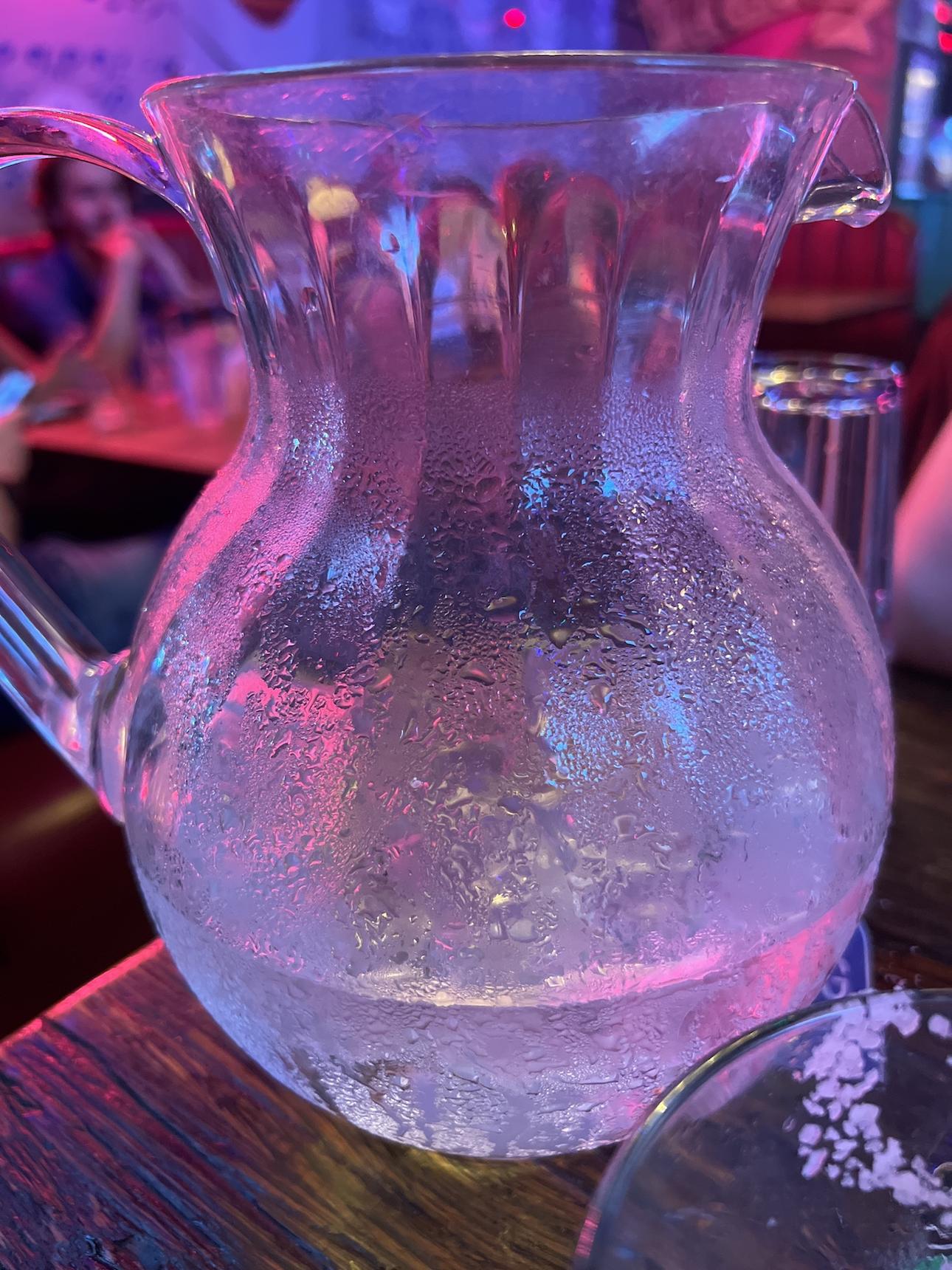 A jug of water and ice colored with pinks and purples from the mood lights